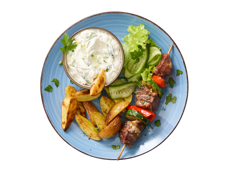 Mesno-zelenjavni ražnjič, pečen krompir in tzatziki na modrem krožniku.