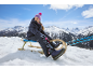 Otrok v smučarski obleki in kapi, sedi na lesenih saneh v zasneženi gorski pokrajini.