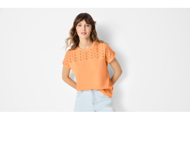 Ženska v oranžnem topu s kratkimi rokavi z vezenino in svetlo modrimi jeans kratkimi hlačami.