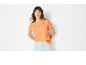 Ženska v oranžnem topu s kratkimi rokavi z vezenino in svetlo modrimi jeans kratkimi hlačami.