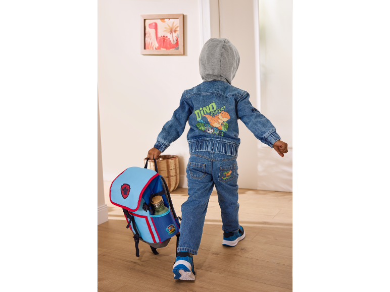 Deček v jeans jakni z dinozavri in jeans hlačah, ki nosi moder nahrbtnik.