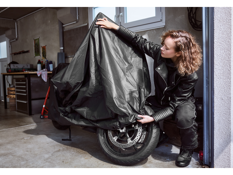 Ženska v usnjeni opremi pokriva motorno kolo s črno zaščitno prevleko v garaži.