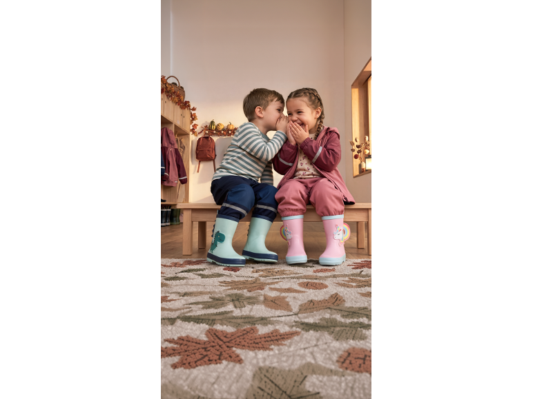 Otroka sedita na klopi, obuta v gumijaste škornje z dinozavrom in samorogom, v jesenskem okolju.