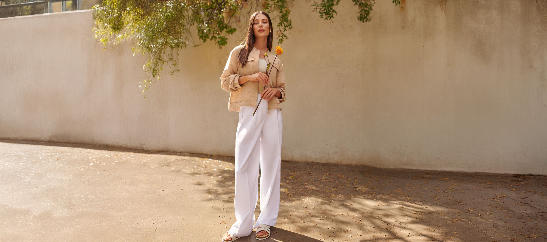 Ženska v bež jakni, belih hlačah in sandalih, drži rumen cvet.