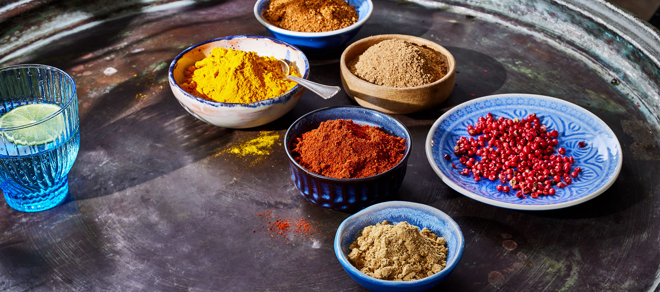 Različne začimbe, kot so kurkuma, paprika in rožnati poper, v skledah na temni površini.