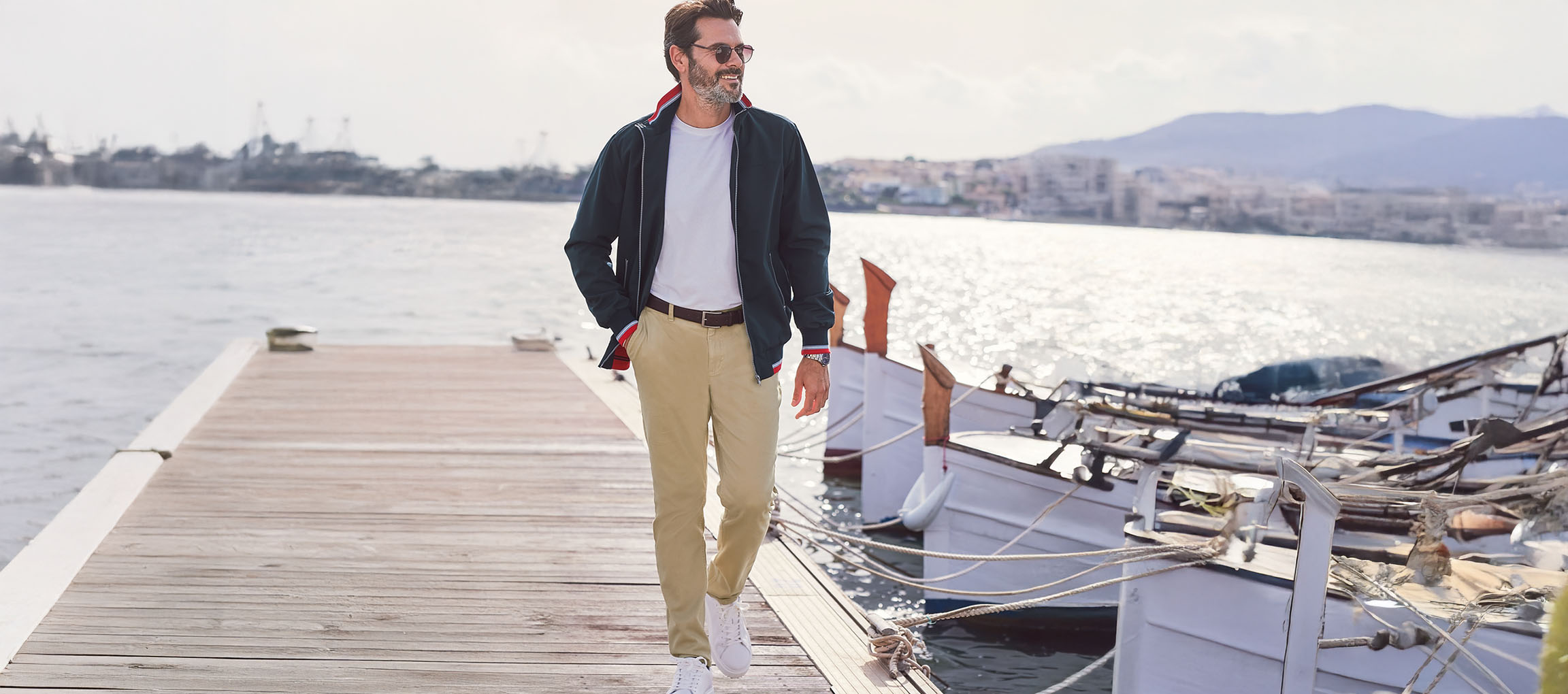 Moški v temno zeleni bomber jakni, chino hlačah in belih supergah na pomolu.
