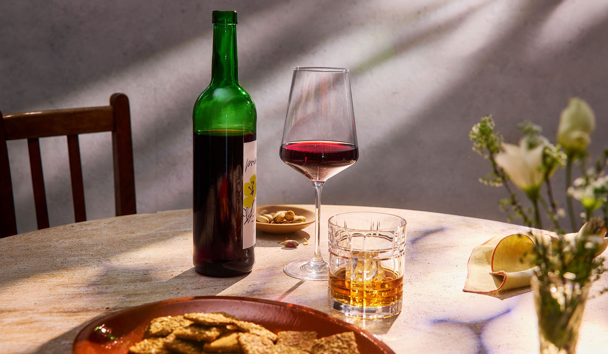 Rdeče vino, viski in prigrizki na mizi v sončni svetlobi.