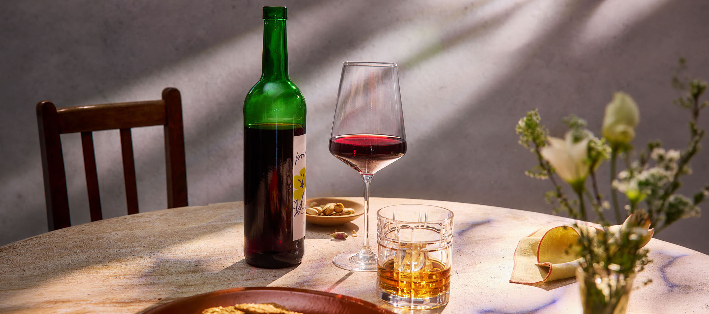 Steklenica rdečega vina, kozarec vina in viski na mizi, s stolom in rožami v ozadju.