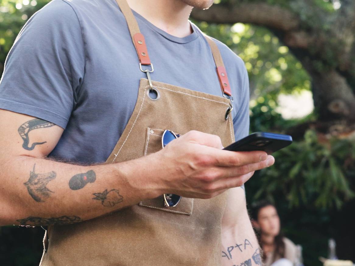 Moški v predpasniku in majici uporablja telefon, v ozadju je drevo.