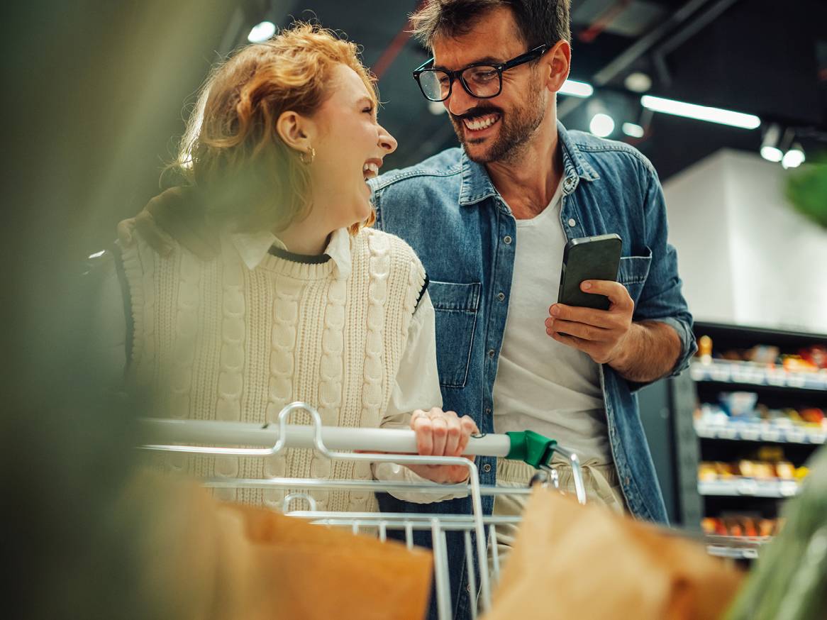 Nasmejan par v trgovini, ženska potiska voziček, moški drži telefon.
