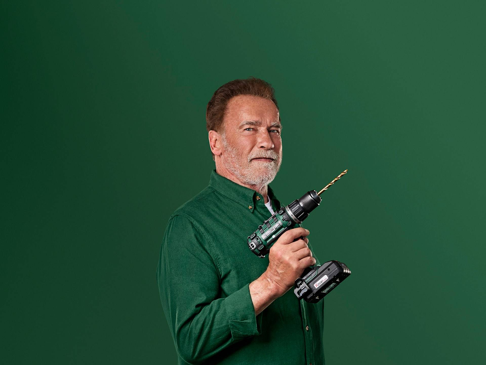 Arnold Schwarzenegger drži zeleni električni vrtalnik na zelenem ozadju.