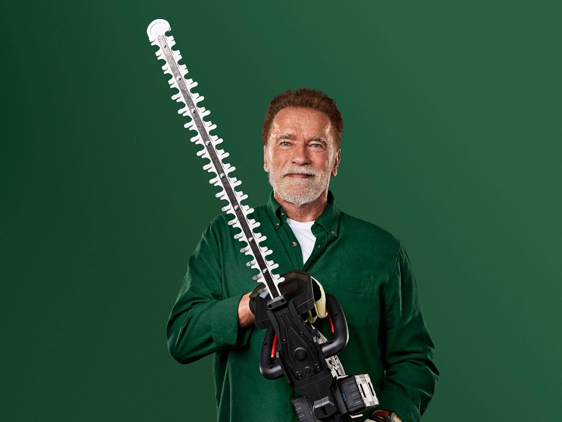 Arnold Schwarzenegger drži velike škarje za živo mejo na zelenem ozadju.