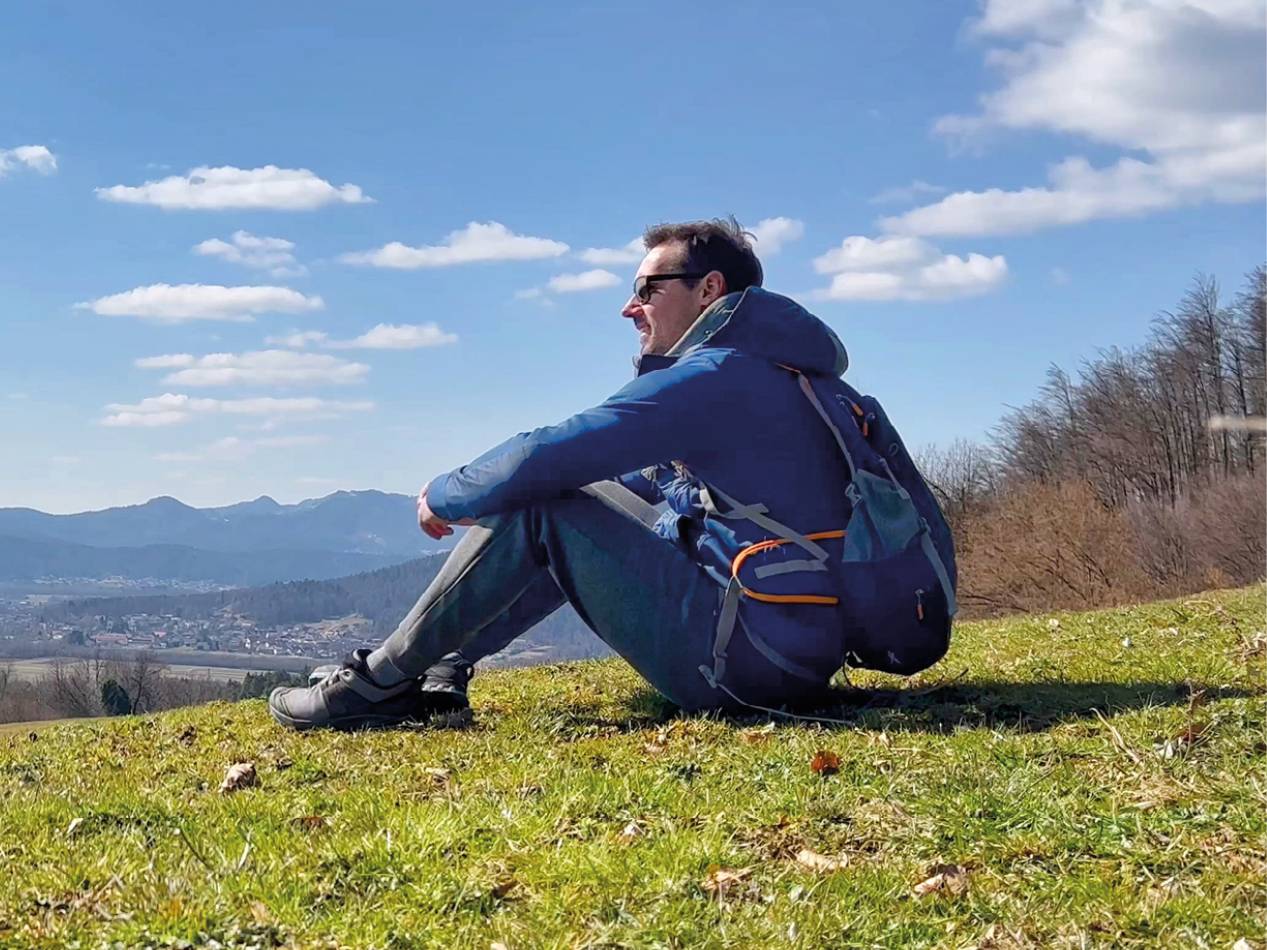 Moški v modri jakni in s pohodniškim nahrbtnikom sedi na travnatem hribu in gleda na pokrajino.