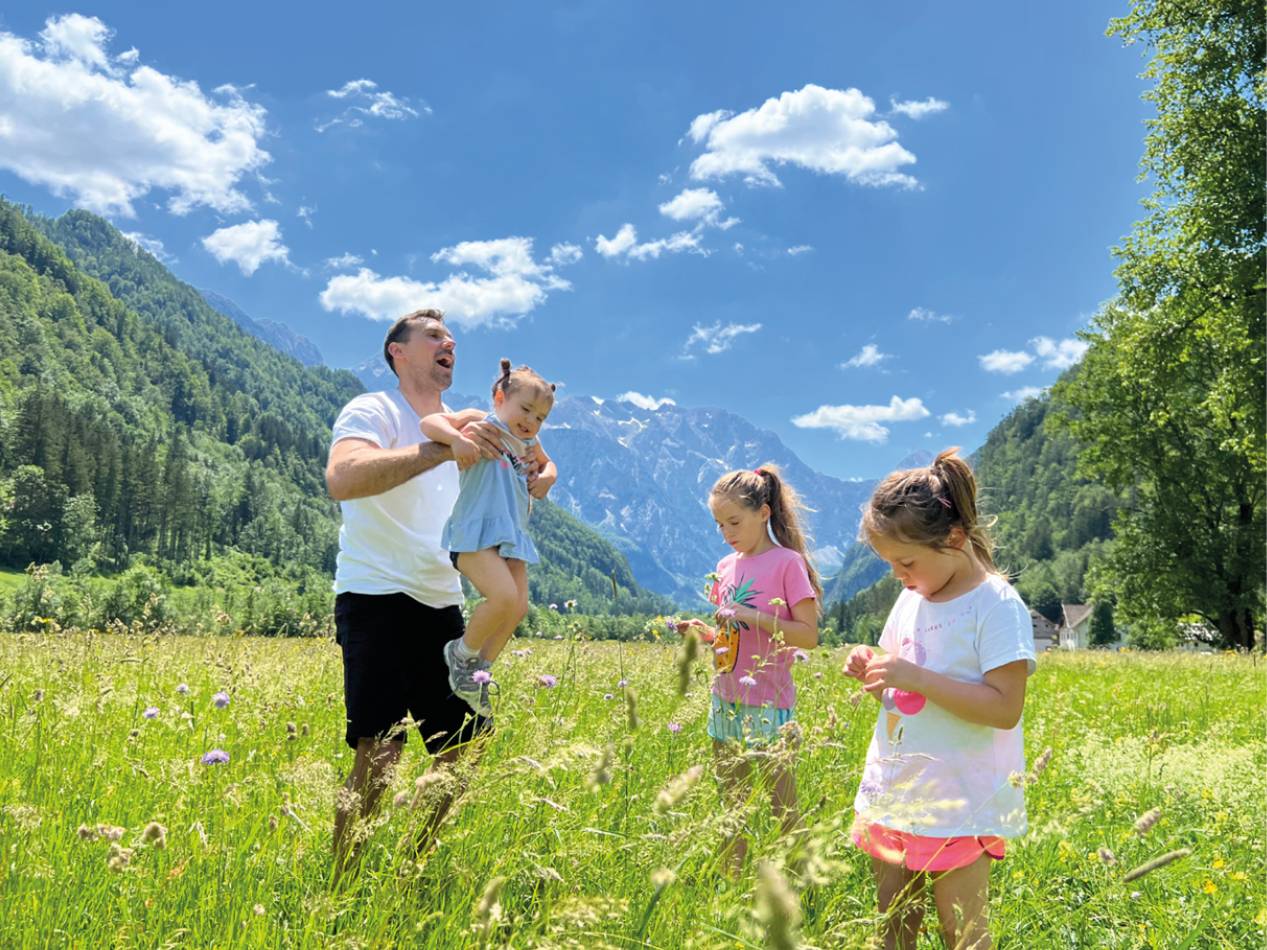 Družina v gorah, oče drži deklico, drugi dve deklici nabirata rože.
