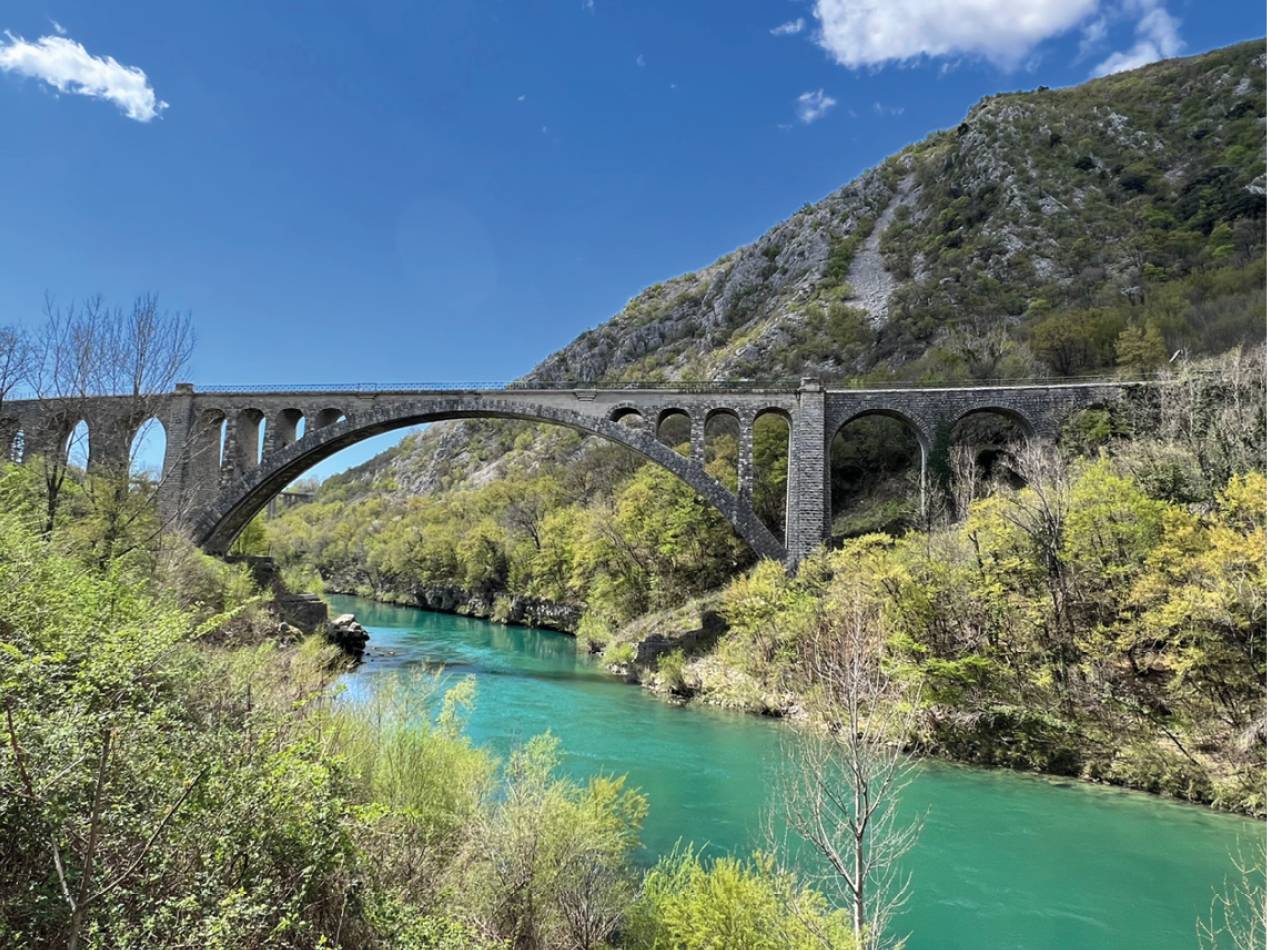 Kamniti most z loki prečka reko s turkizno vodo, obdan z zelenimi drevesi in gorami.