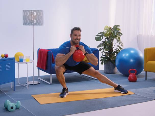 Moški izvaja stranski izpad z rdečo utežjo kettlebell v dnevni sobi.