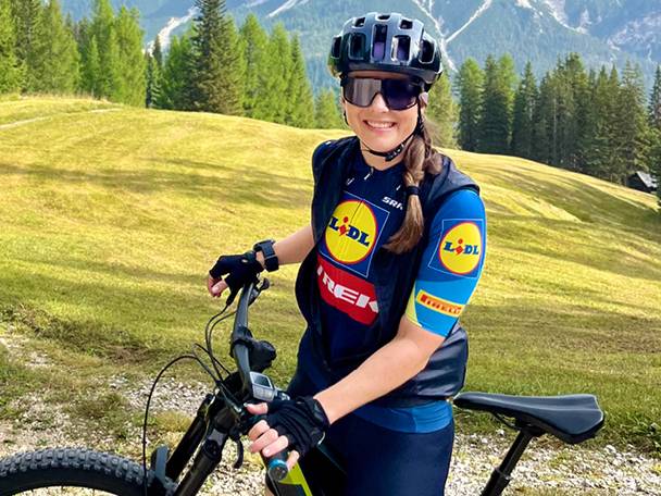 Ženska v kolesarski opremi Lidl in Trek, s čelado in sončnimi očali, stoji ob gorskem kolesu.