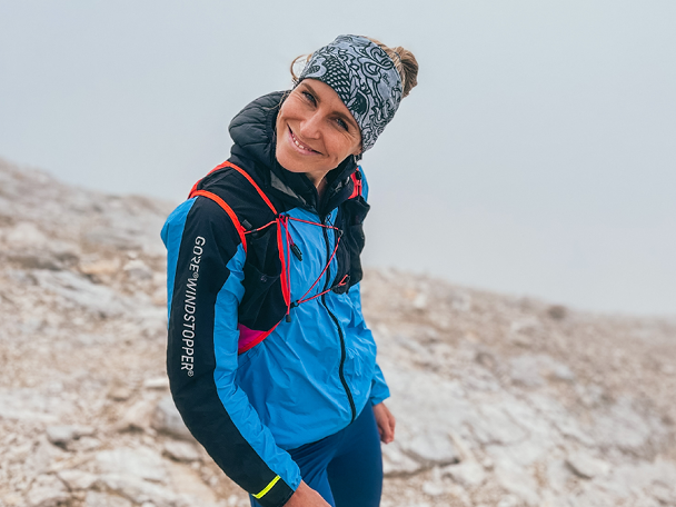 Ženska v modri jakni Gore Windstopper in naglavnem traku se nasmehne na skalnati gori.