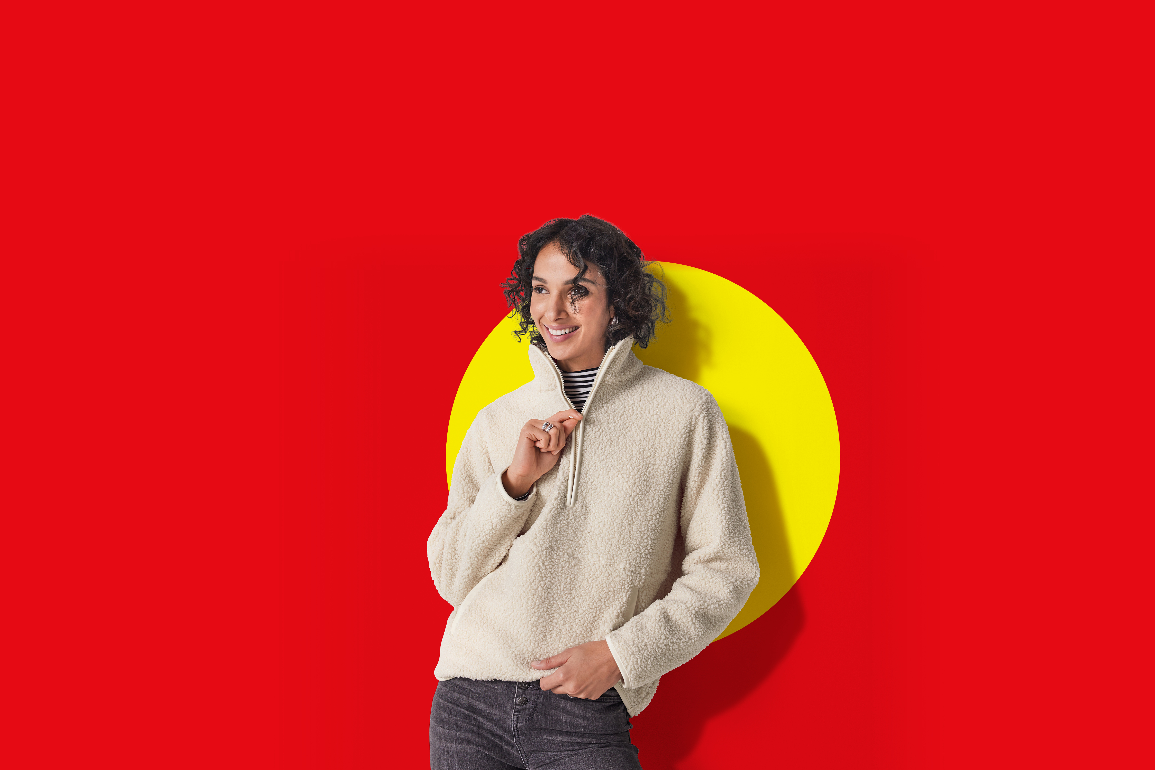 Ženska v bež puloverju iz flisa in črnih kavbojkah, nasmejana, na rdečem ozadju z rumenim krogom.