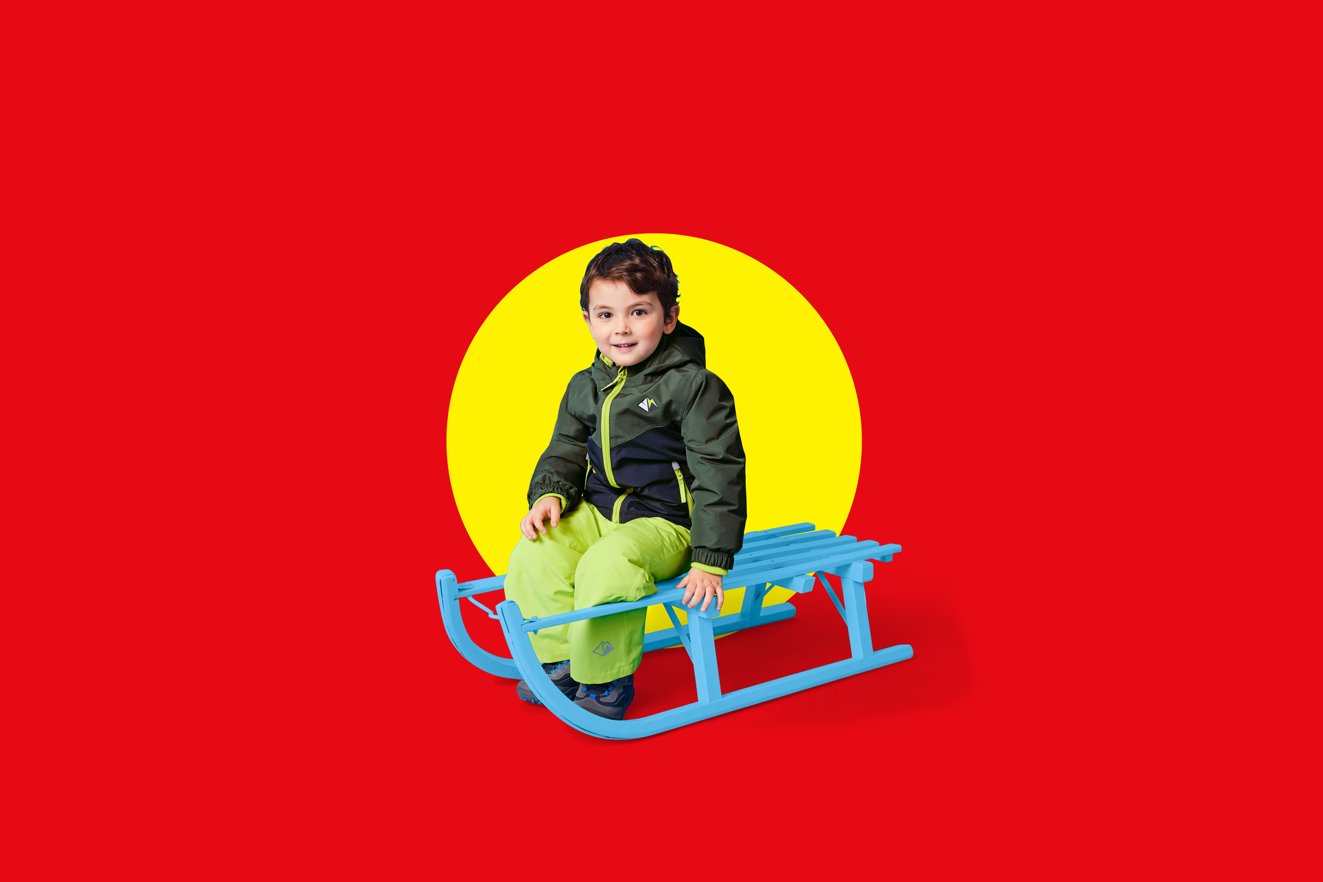 Deček v zimski jakni in hlačah sedi na modrih saneh pred rdečim ozadjem z rumenim krogom.