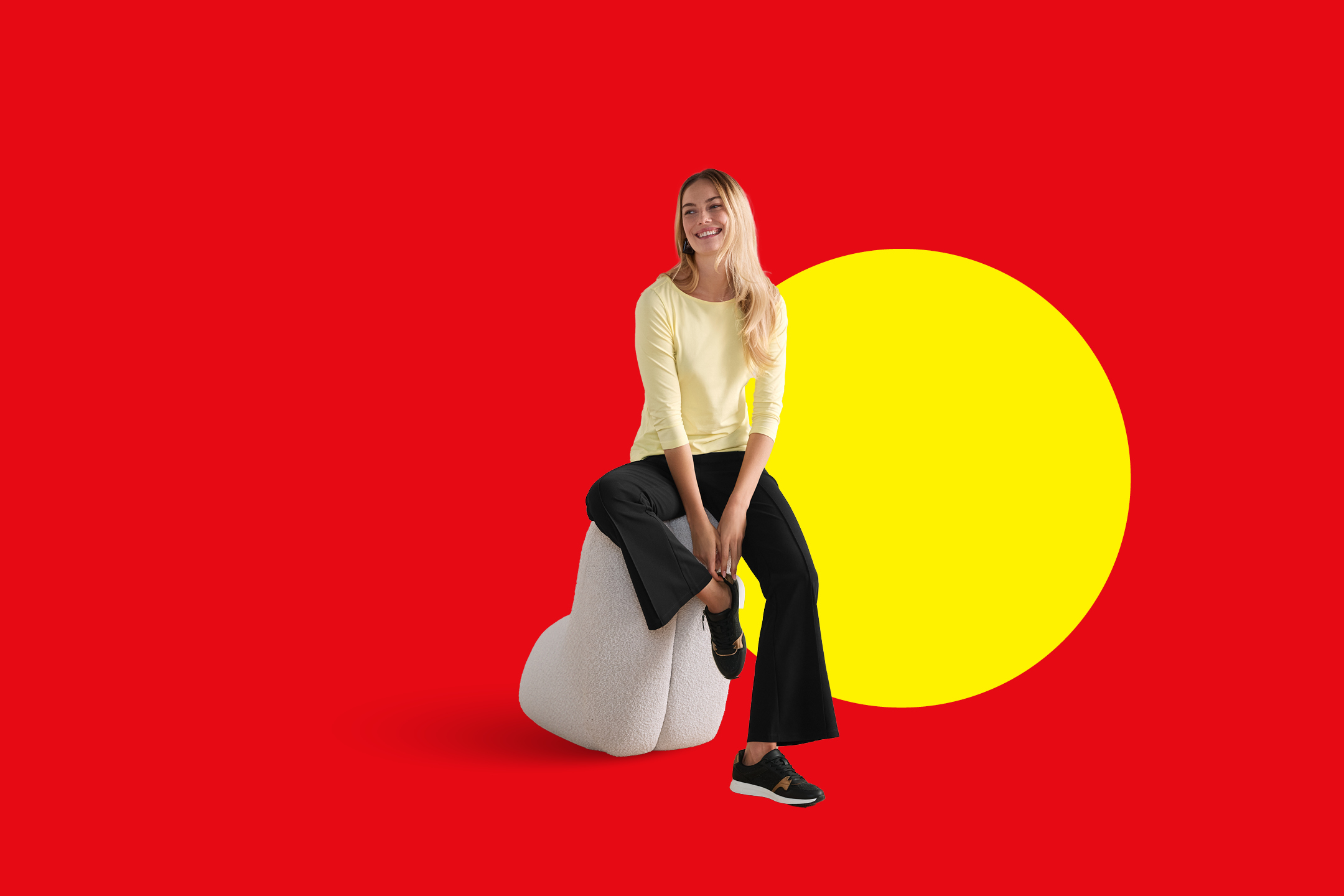 Ženska v svetlo rumeni majici in črnih hlačah sedi na beli blazini v obliki srca.