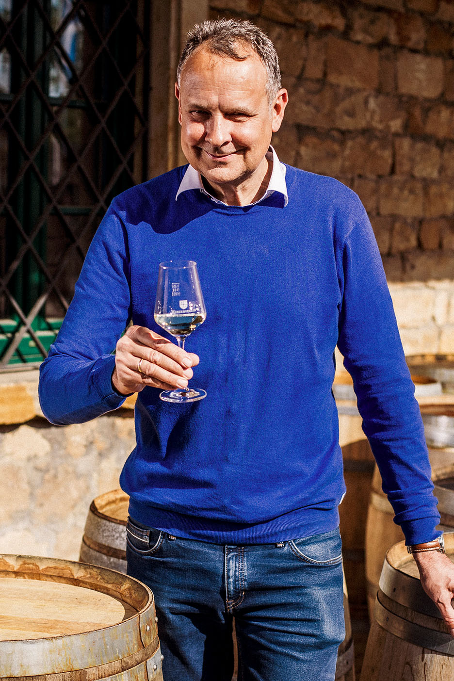 Nasmejan moški v modrem puloverju in kavbojkah drži kozarec belega vina med lesenimi sodi.
