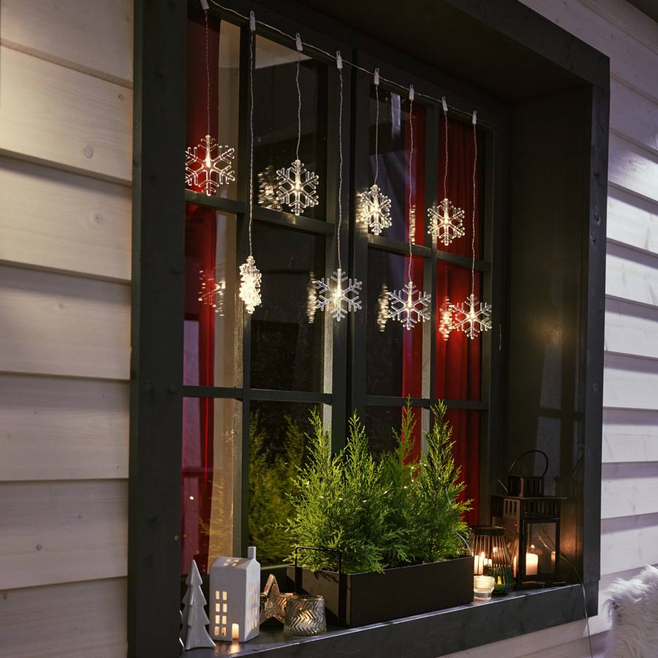 Svetlobna veriga snežink visi v oknu z majhnimi jelkami in svečami na okenski polici.