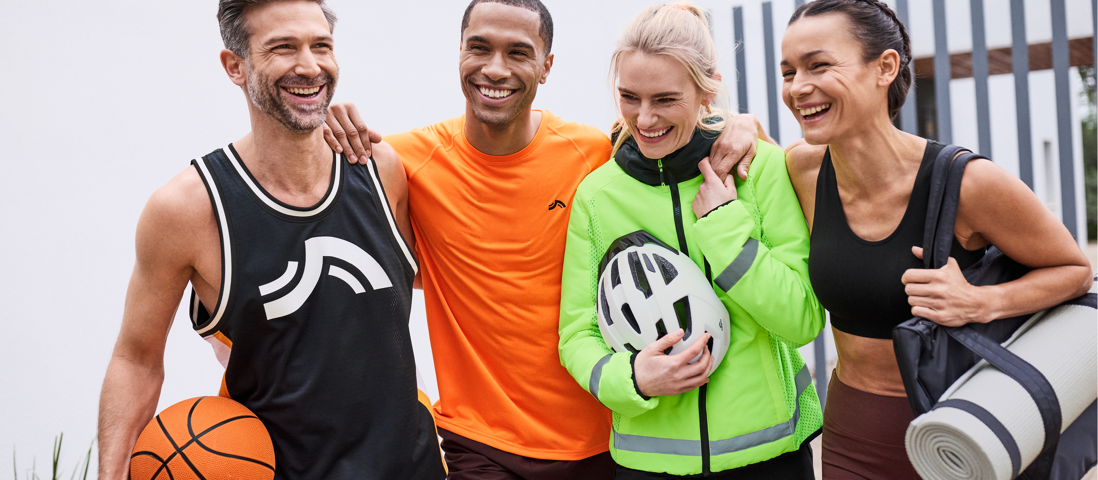 Štirje nasmejani prijatelji v športnih oblačilih, vključno s kolesarsko jakno in košarkarsko majico.
