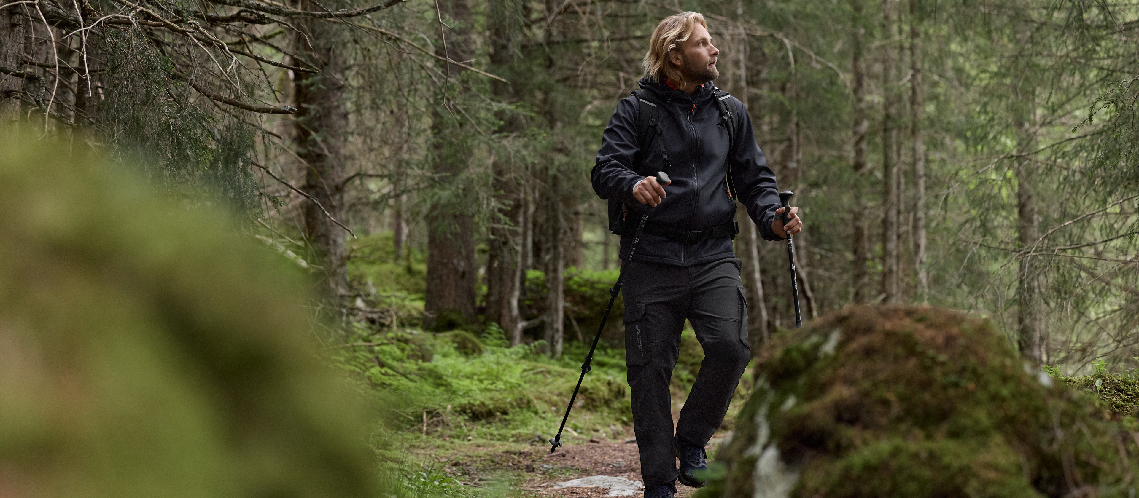 Moški pohodnik v črni jakni in hlačah s pohodniškimi palicami v gozdu.