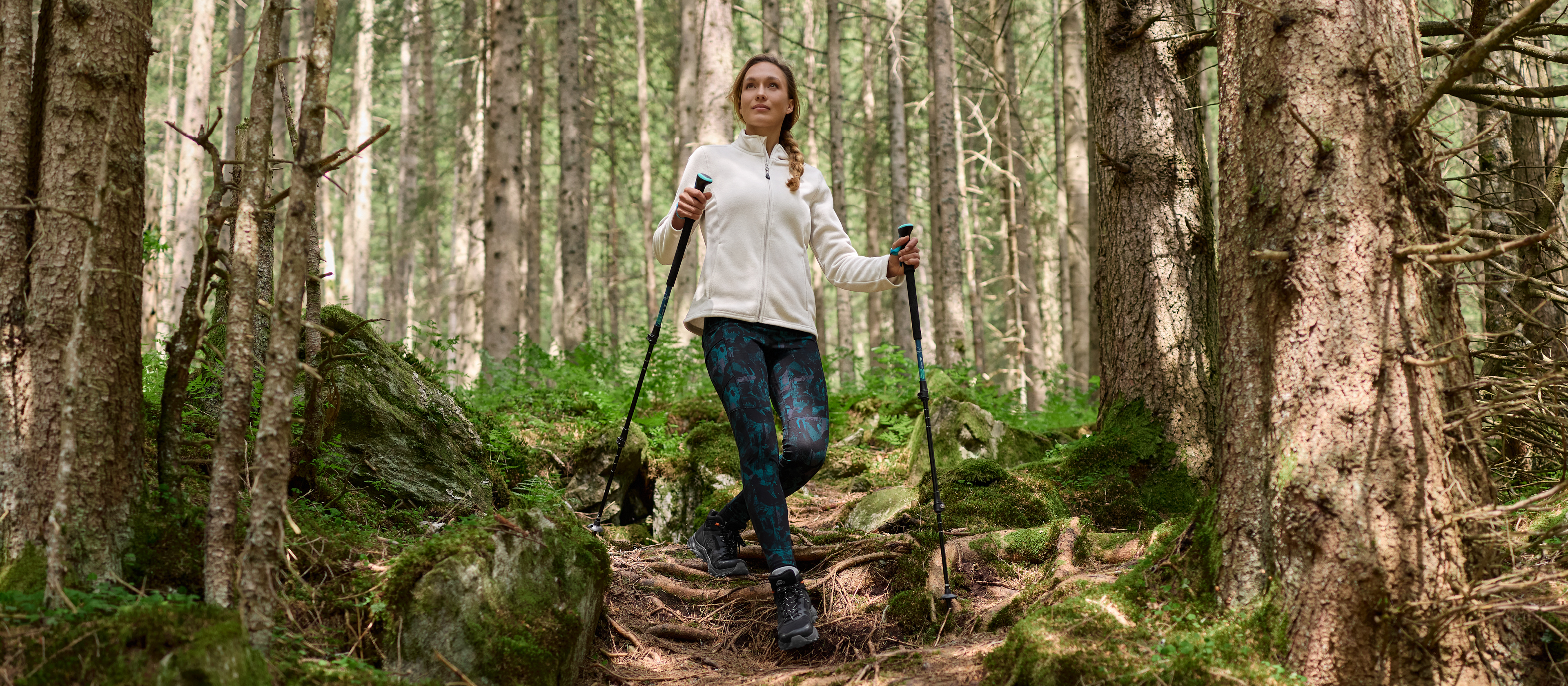 Ženska v belem flisu in vzorčastih pajkicah hodi po gozdni poti s palicami.