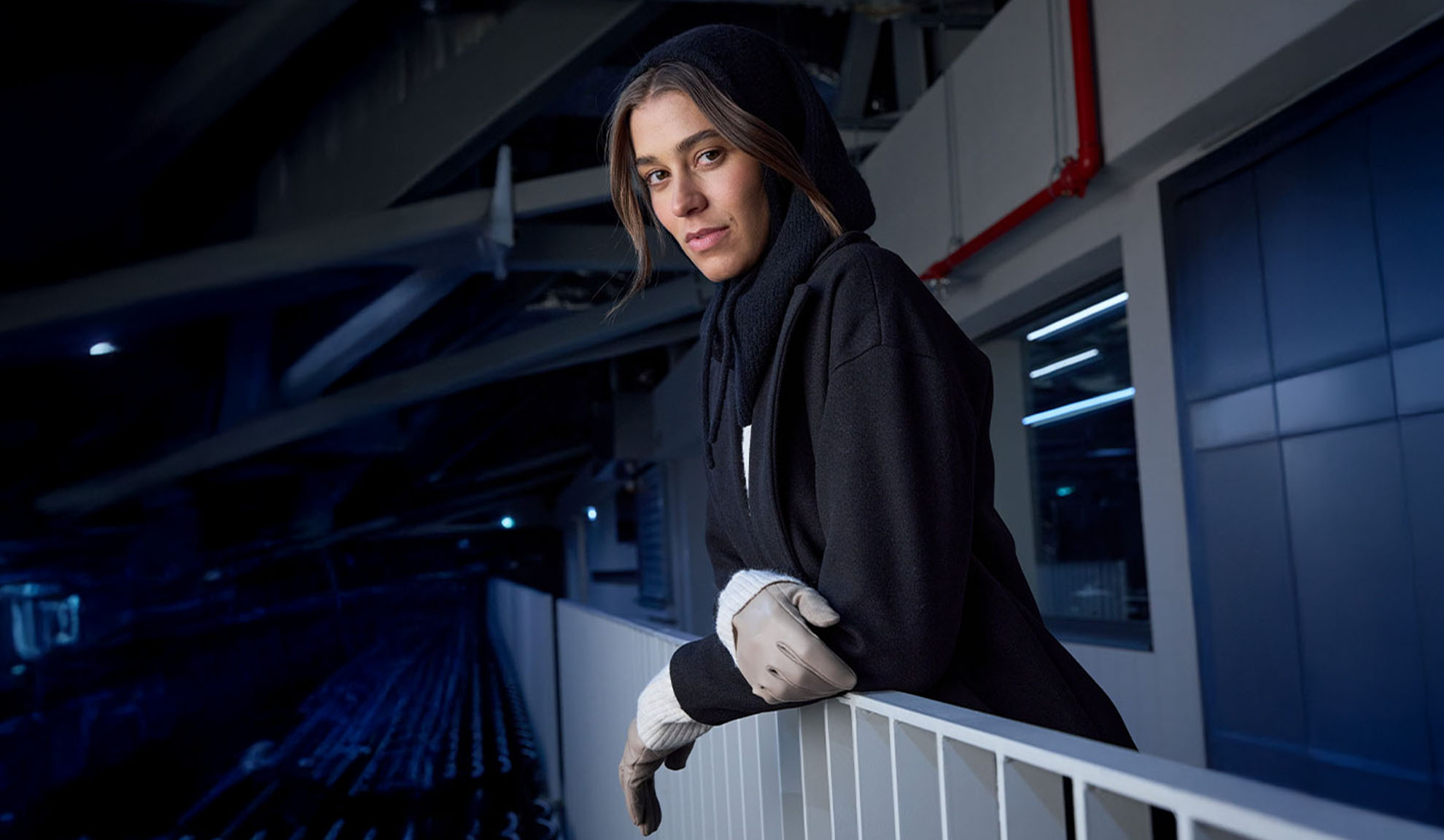 Ženska v črnem plašču in kapuci, naslonjena na ograjo v modernem okolju.