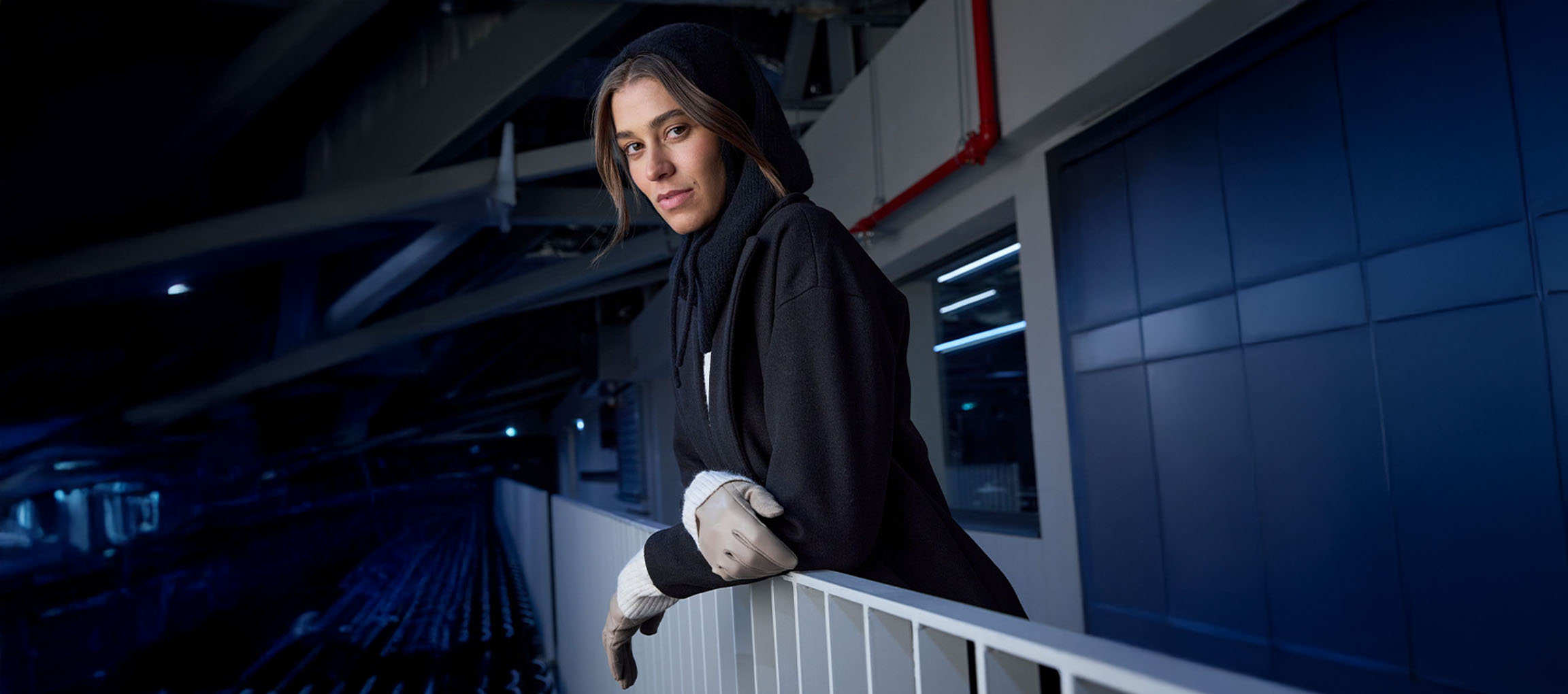 Ženska v črnem plašču in rokavicah naslonjena na ograjo v urbanem okolju.