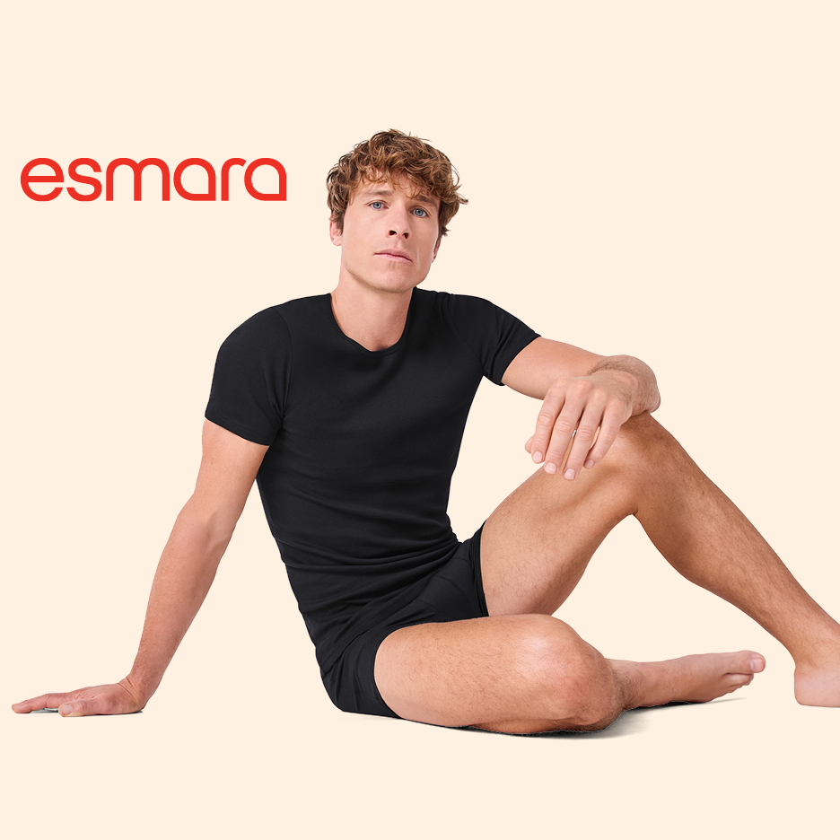 Moški v črni majici in boksaricah sedi na tleh, zgoraj levo rdeč napis esmara.