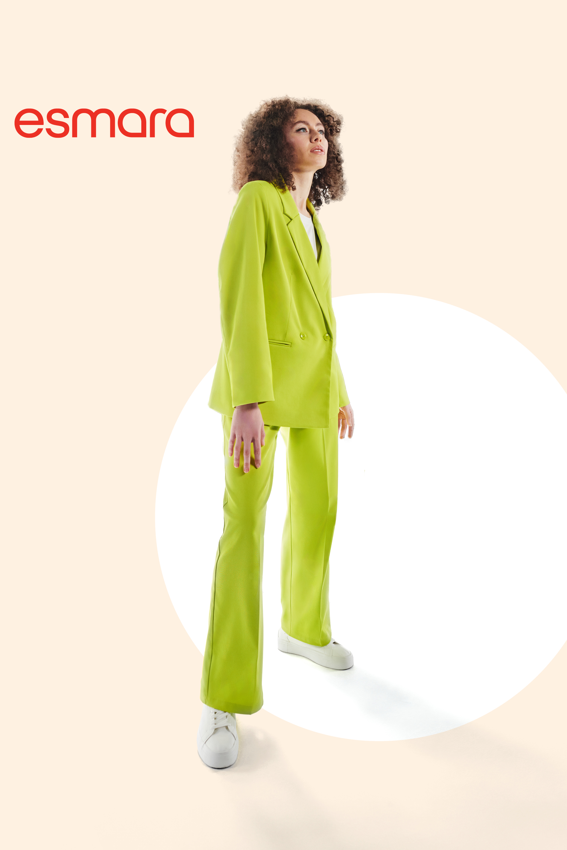 Ženska v limetasto zelenem kostimu s suknjičem in hlačami, belo majico in belimi supergami.