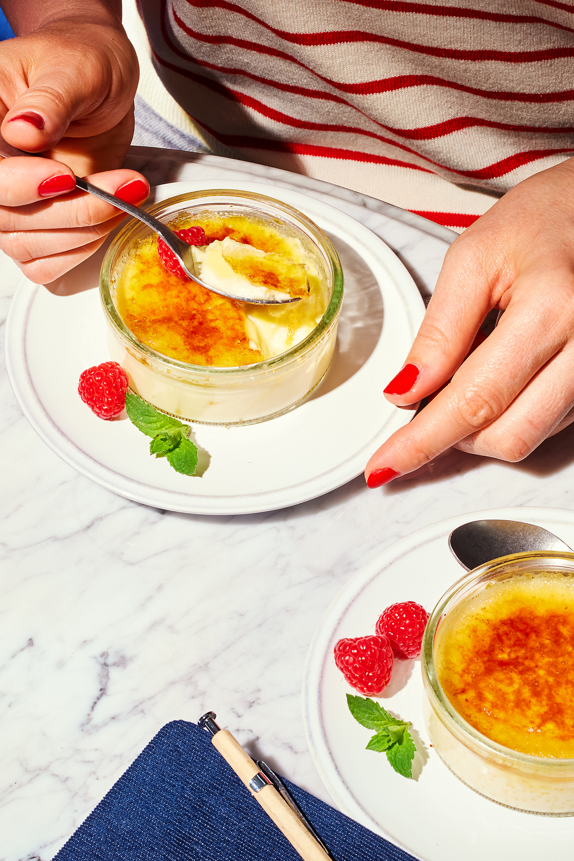 Oseba uživa v crème brûléeju z malinami in meto na marmornati mizi.