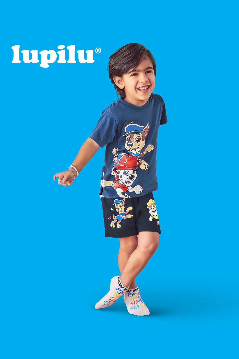 Nasmejan fant v modri majici in kratkih hlačah s potiskom Paw Patrol, belih nogavicah.