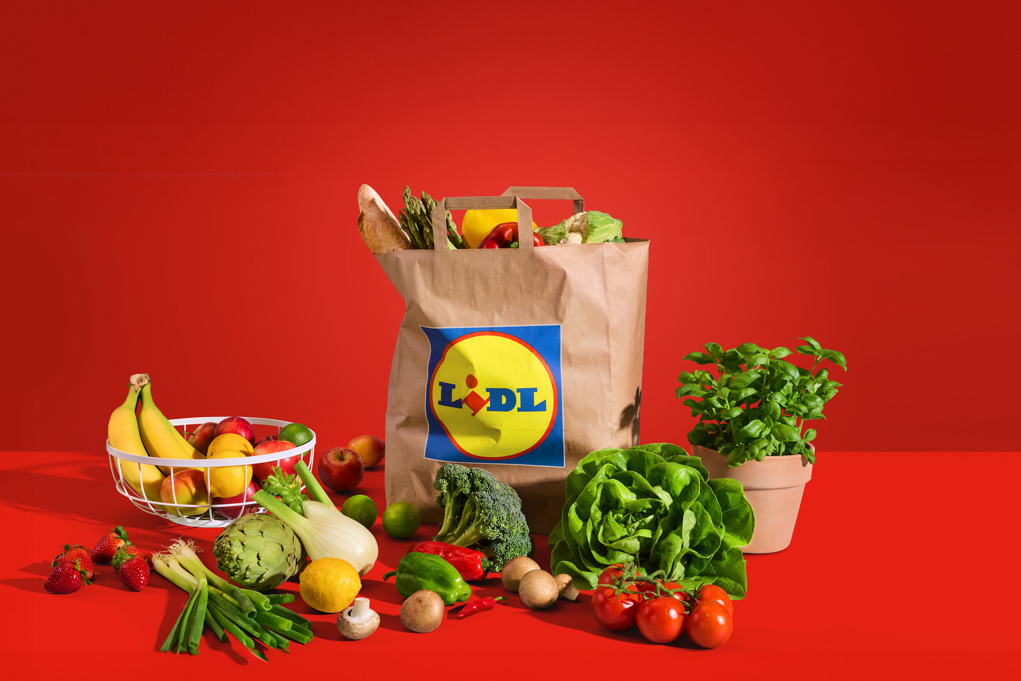 Nakupovalna vrečka s svežo zelenjavo in sadjem na rdečem ozadju.