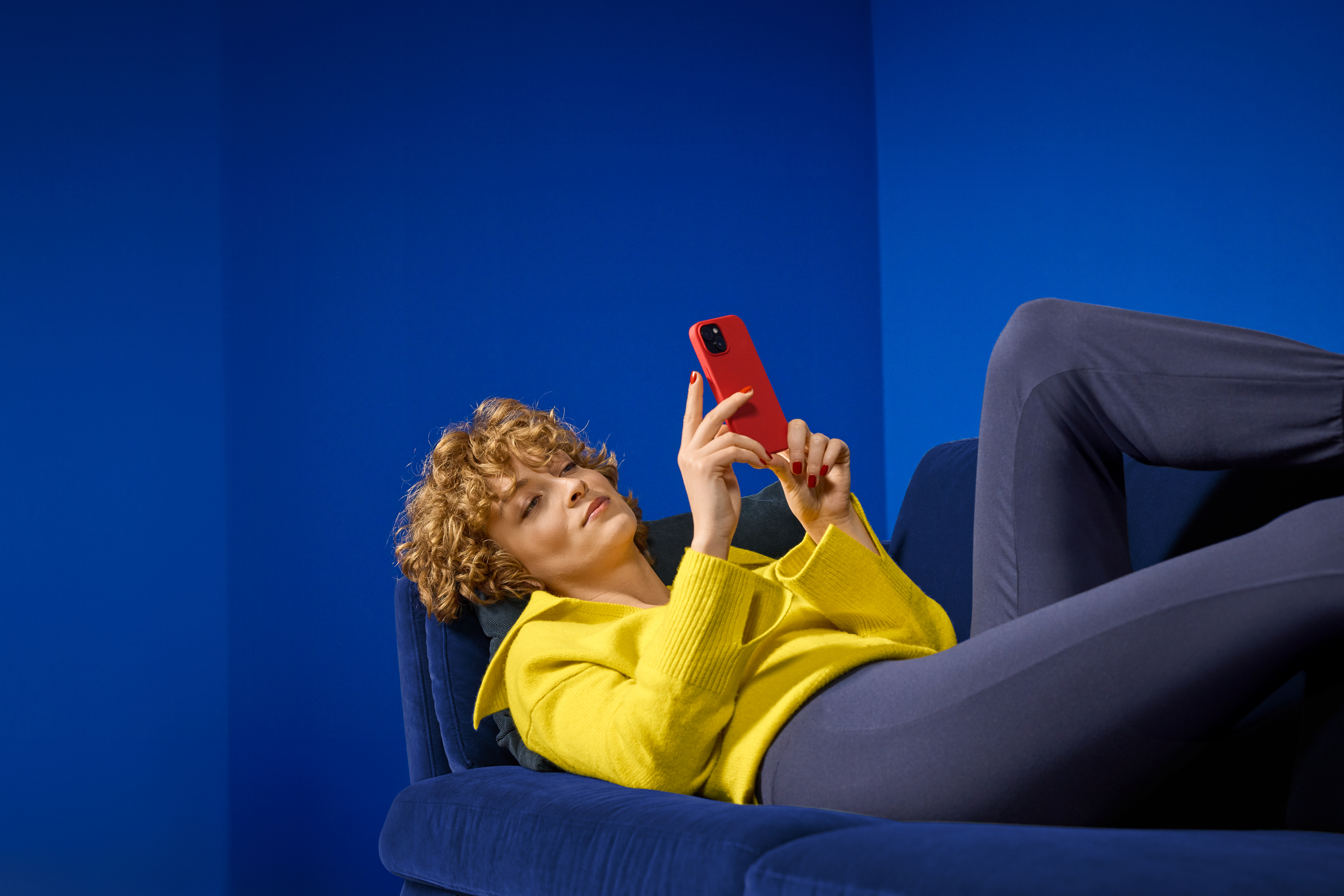 Ženska v rumenemu puloverju in modrih hlačah leži na modri zofi in gleda v rdeč telefon.