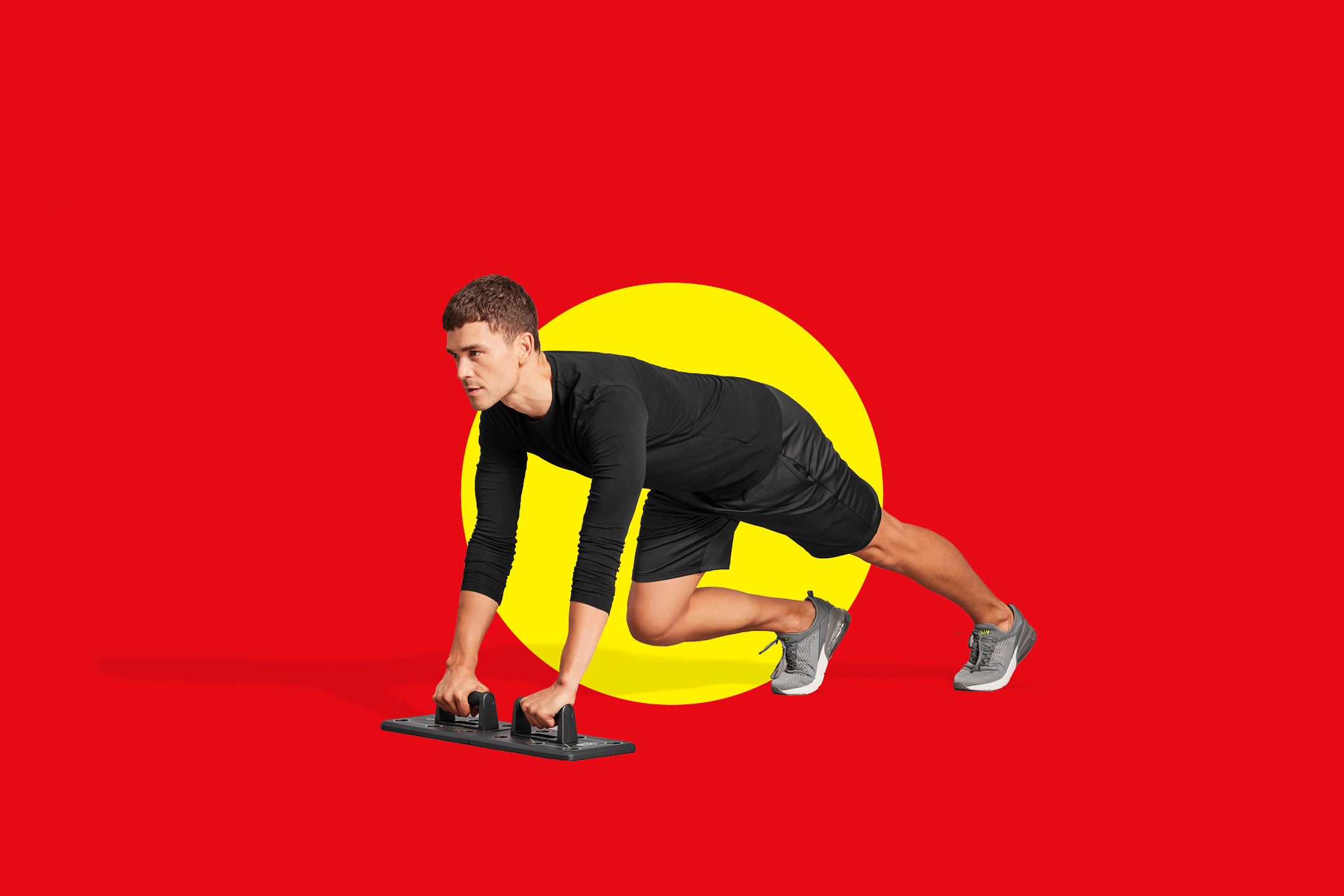 Moški v črni športni opremi dela sklece z ročaji na rdečem ozadju z rumenim krogom.