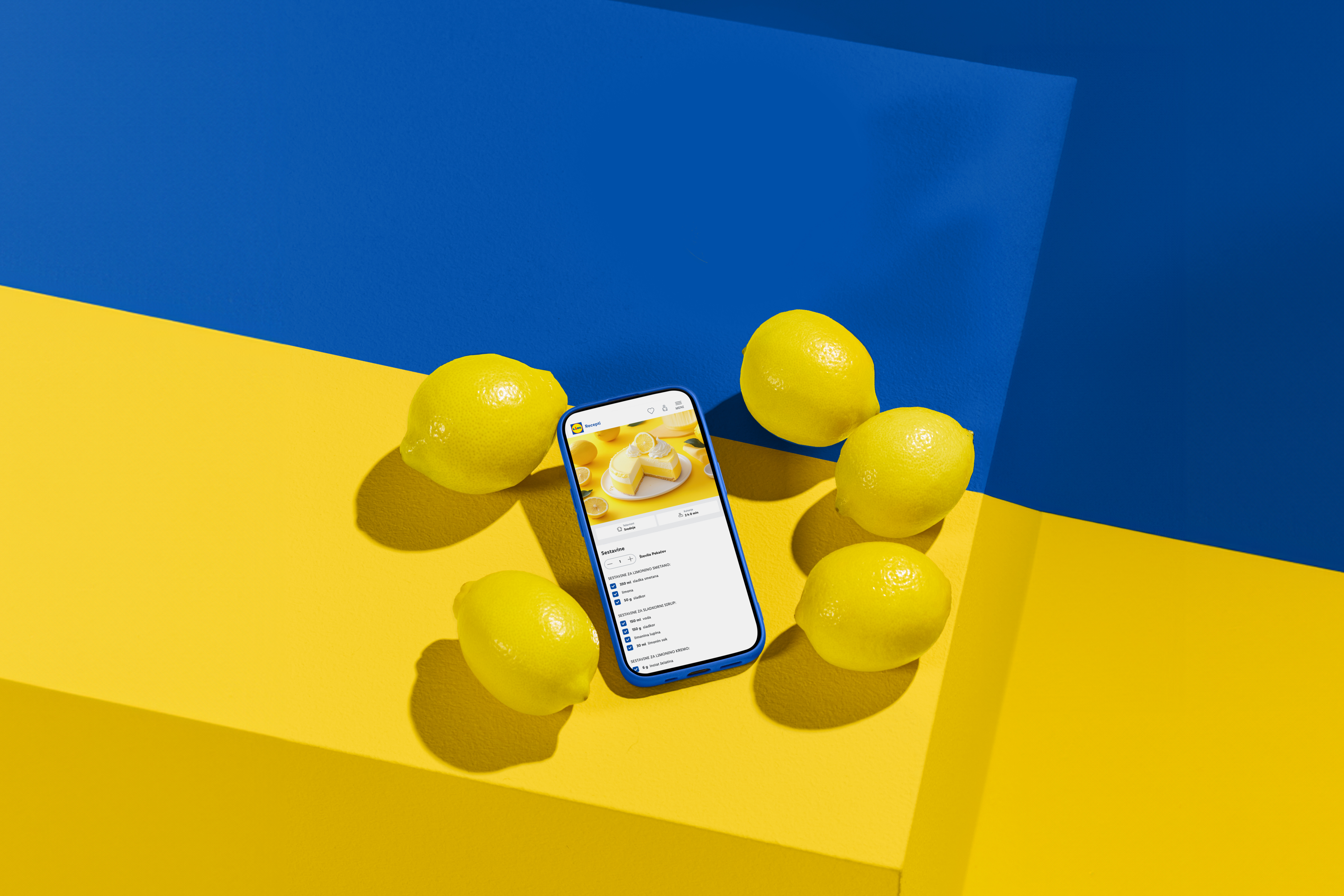 Telefon z receptom za limonino torto in sveže limone na modro-rumenem ozadju.