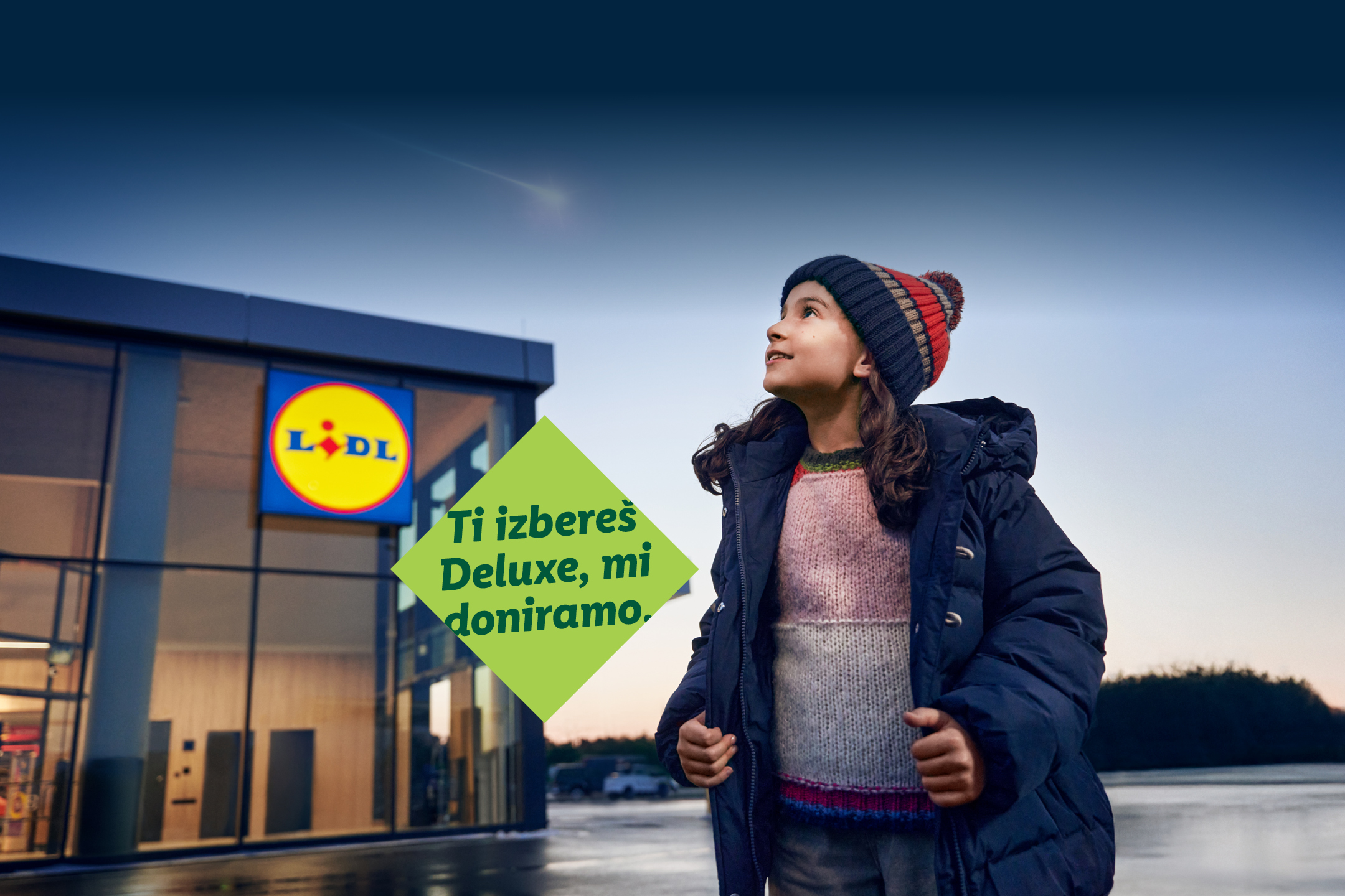 Deklica v zimski obleki pred trgovino Lidl z napisom: »Ti izbereš Deluxe, mi doniramo.«