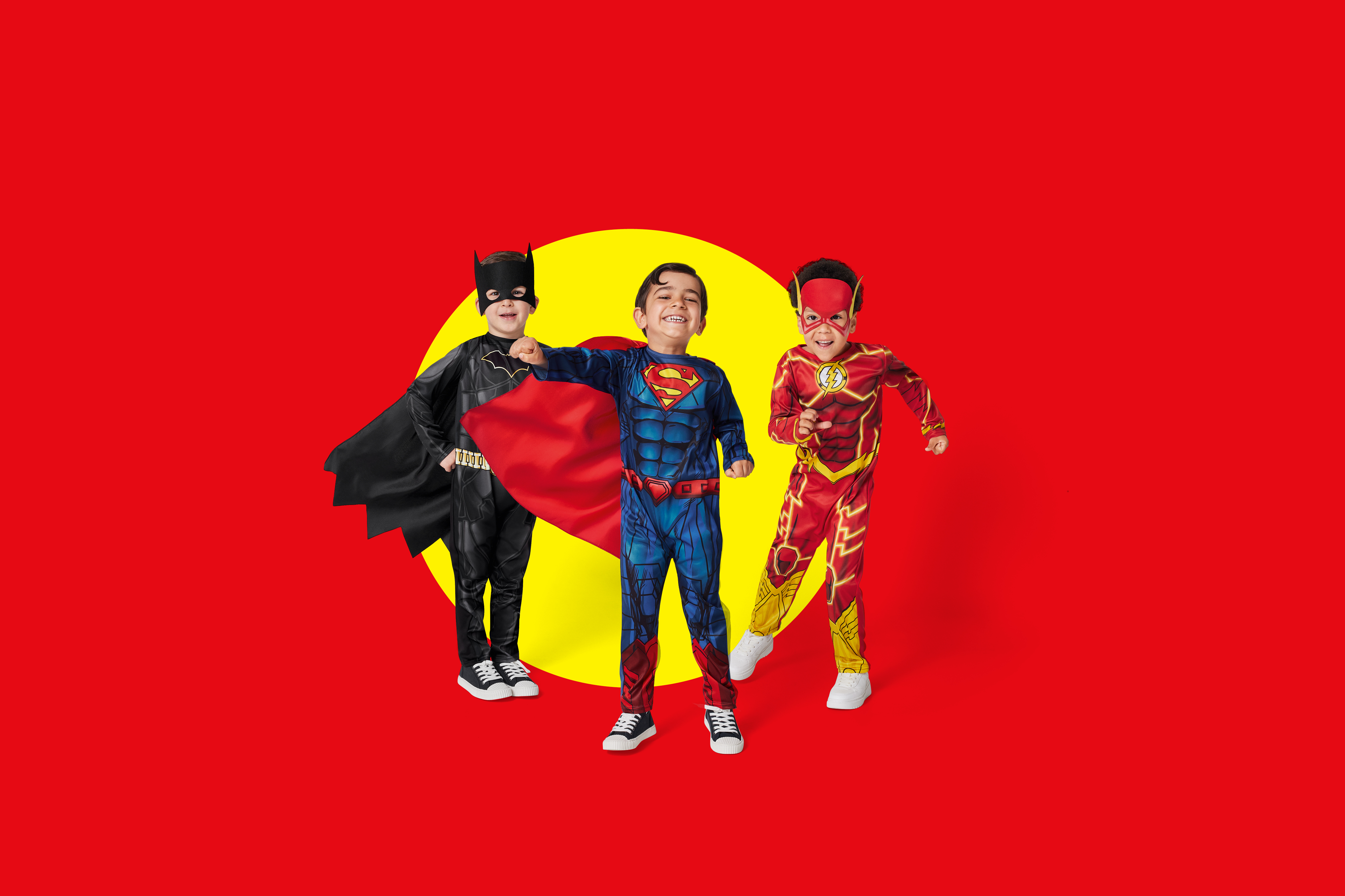 Trije otroci v kostumih Batmana, Supermana in Flasha na rdečem ozadju z rumenim krogom.