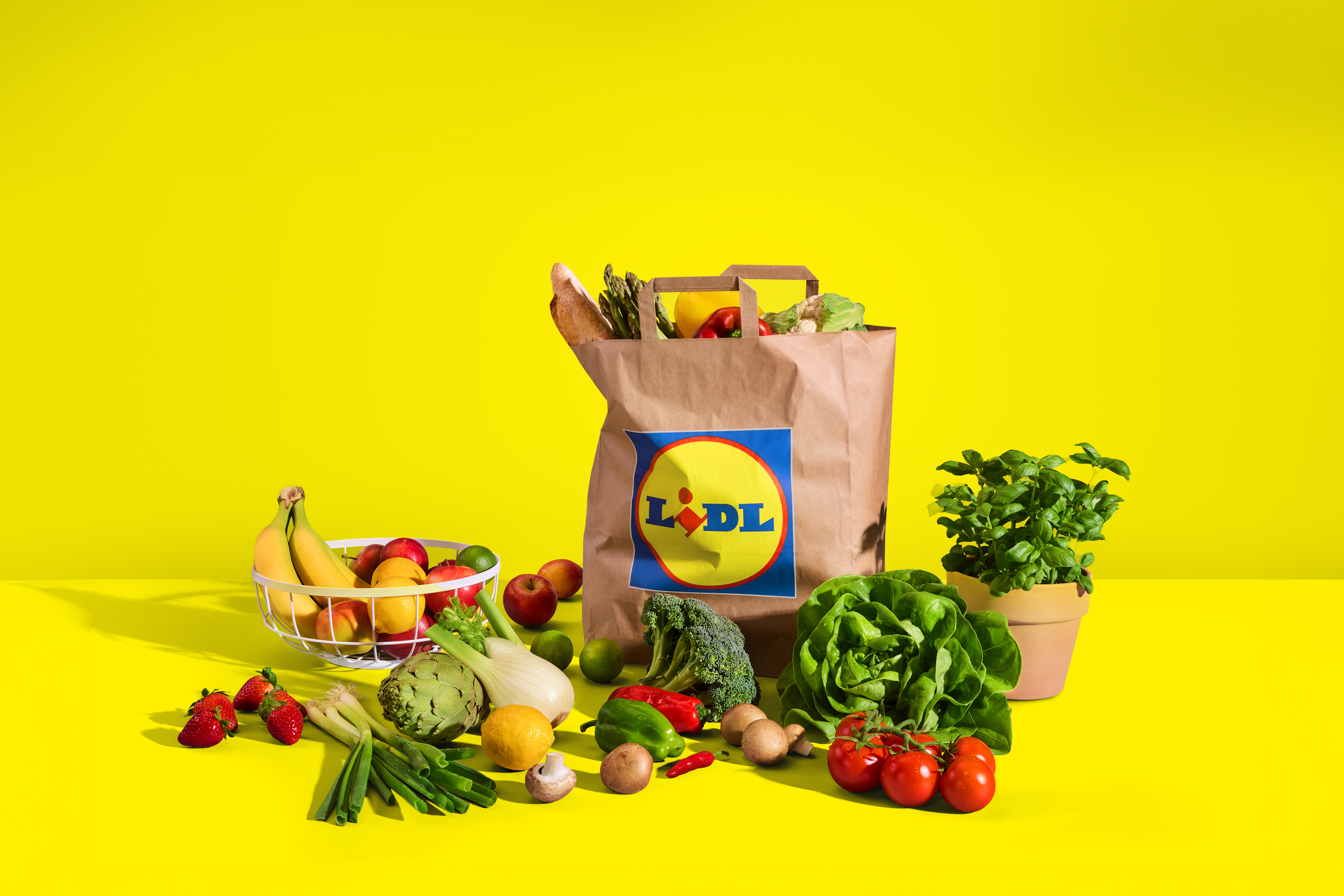 Nakupovalna vrečka s sadjem in zelenjavo, vključno z bananami, solato in paradižnikom, na rumenem ozadju.
