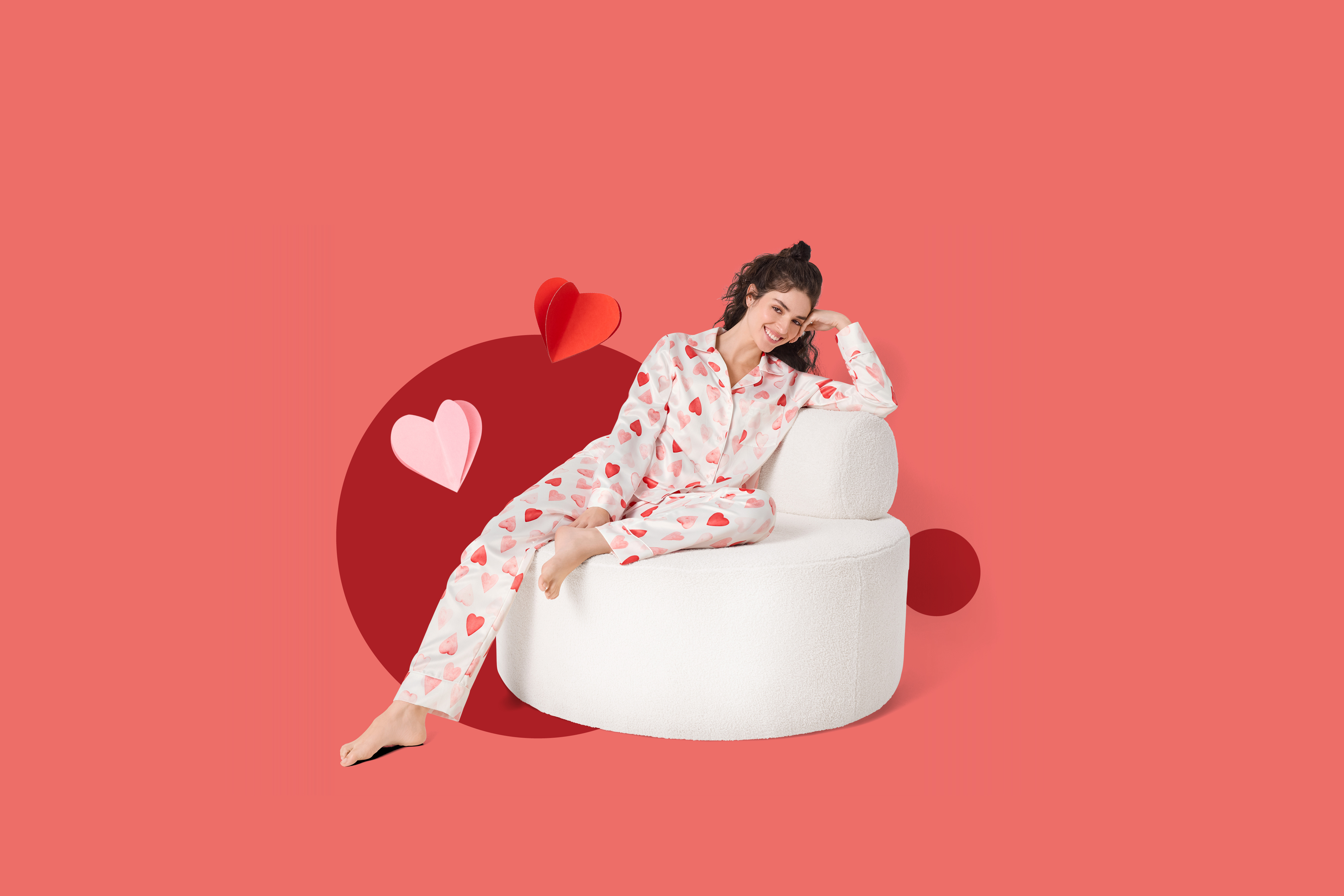 Ženska v pižami s srčki sedi na belem naslanjaču, obkrožena z rdečimi in rožnatimi srčki.