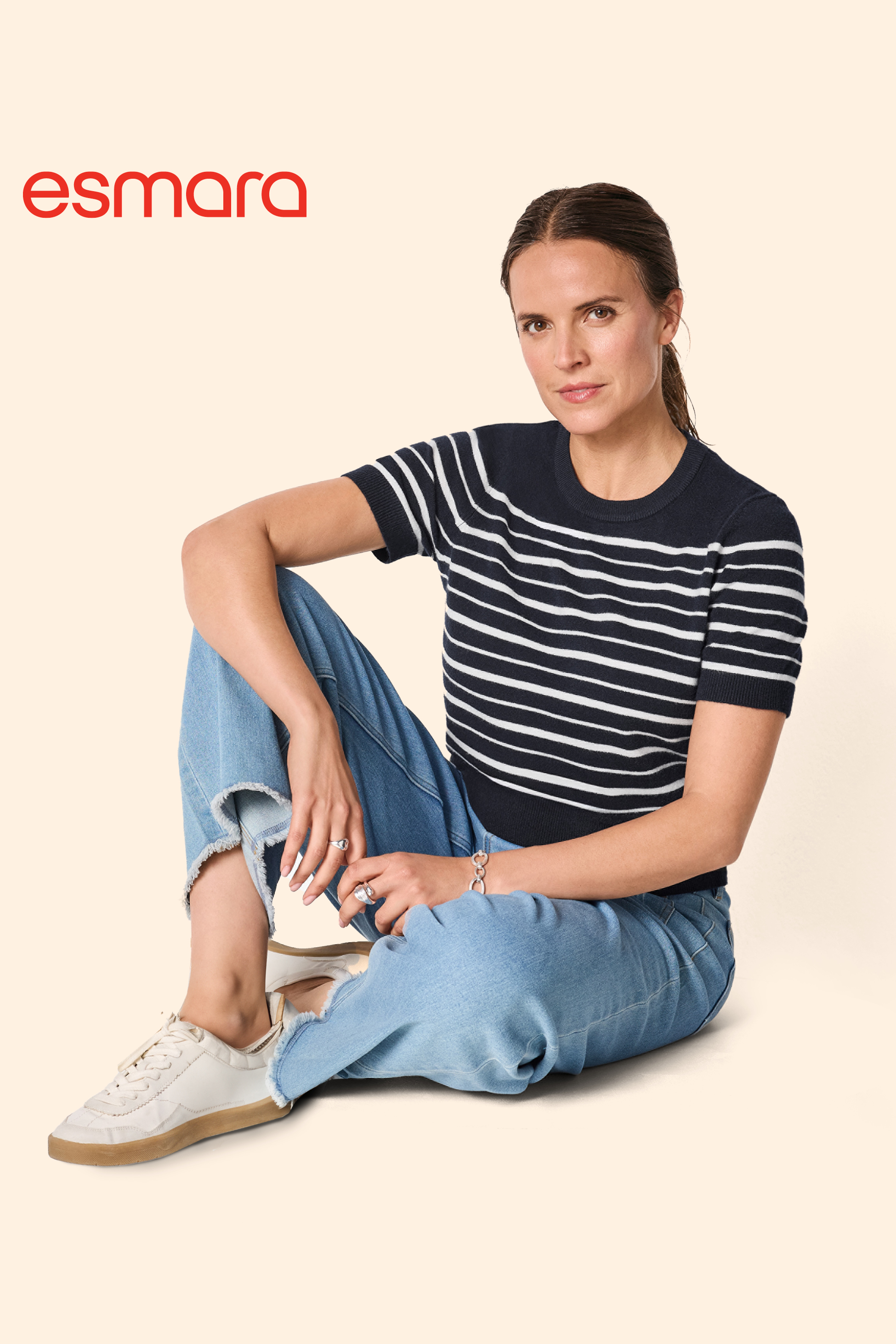 Ženska v črtasti majici in kavbojkah sedi na tleh, z logotipom Esmara v zgornjem levem kotu.