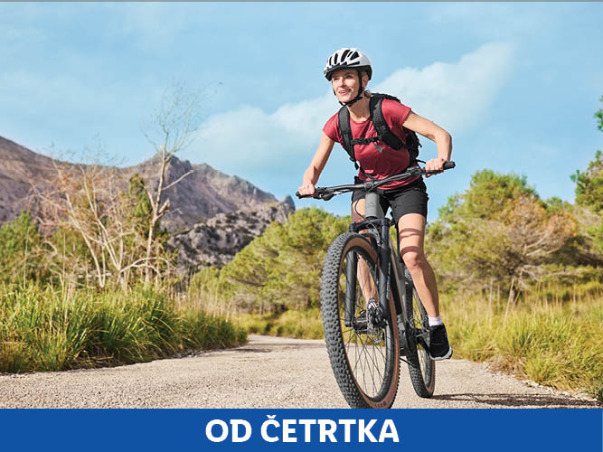 Ženska se vozi s kolesom po makadamski cesti ob gorskem ozadju.