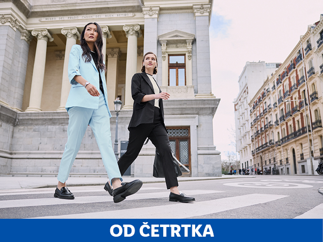 Dve ženski v elegantnih oblekah hodita po prehodu za pešce pred klasično stavbo.
