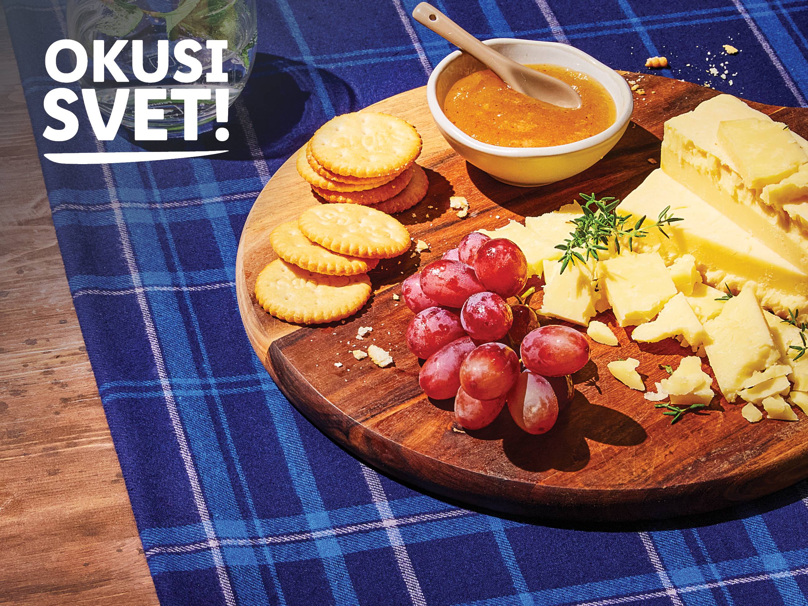 Sir, krekerji, grozdje in marmelada na leseni deski na karirastem prtu.