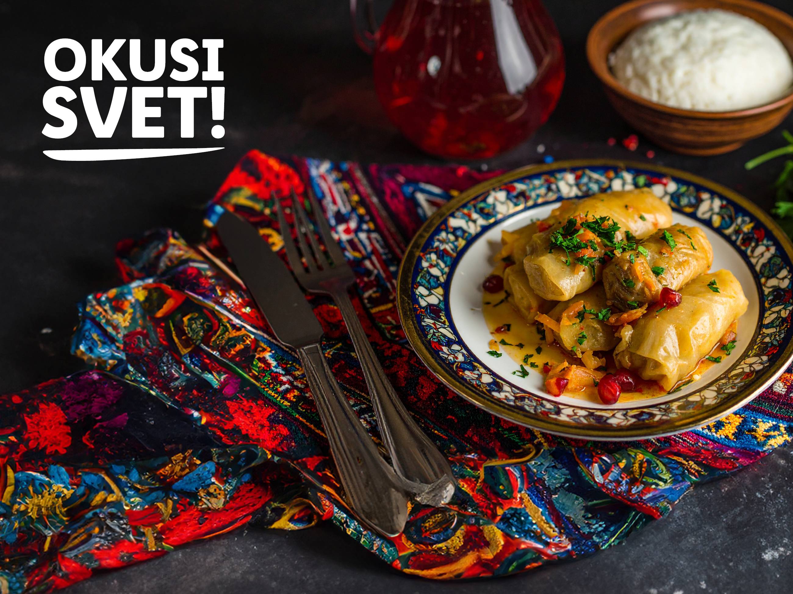 Polnjeni zeljnati zvitki na krožniku z rižem in pijačo, z napisom »OKUSI SVET!«