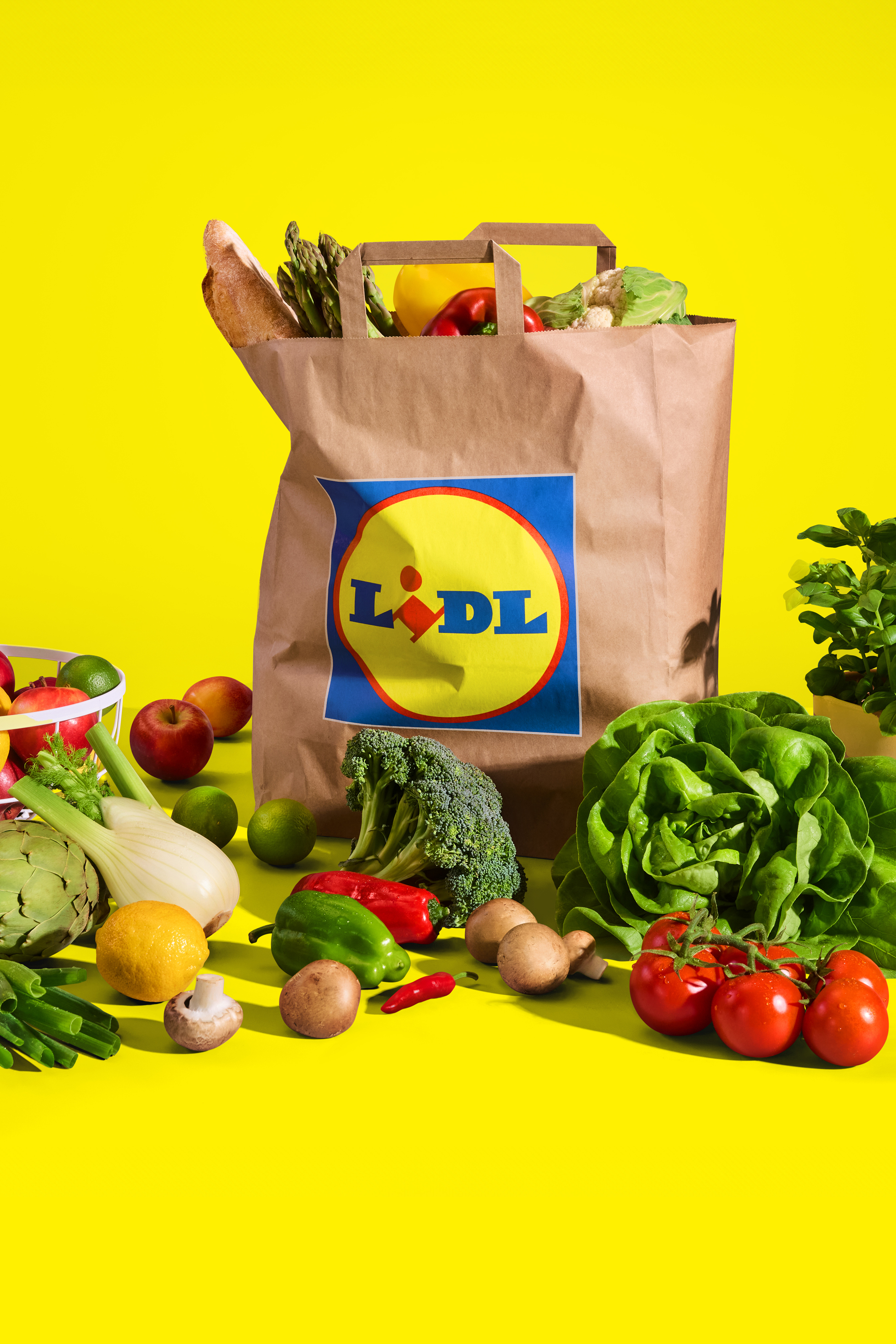 Lidl papirnata vrečka, polna svežih pridelkov, obdana z različnim sadjem in zelenjavo na rumenem ozadju.